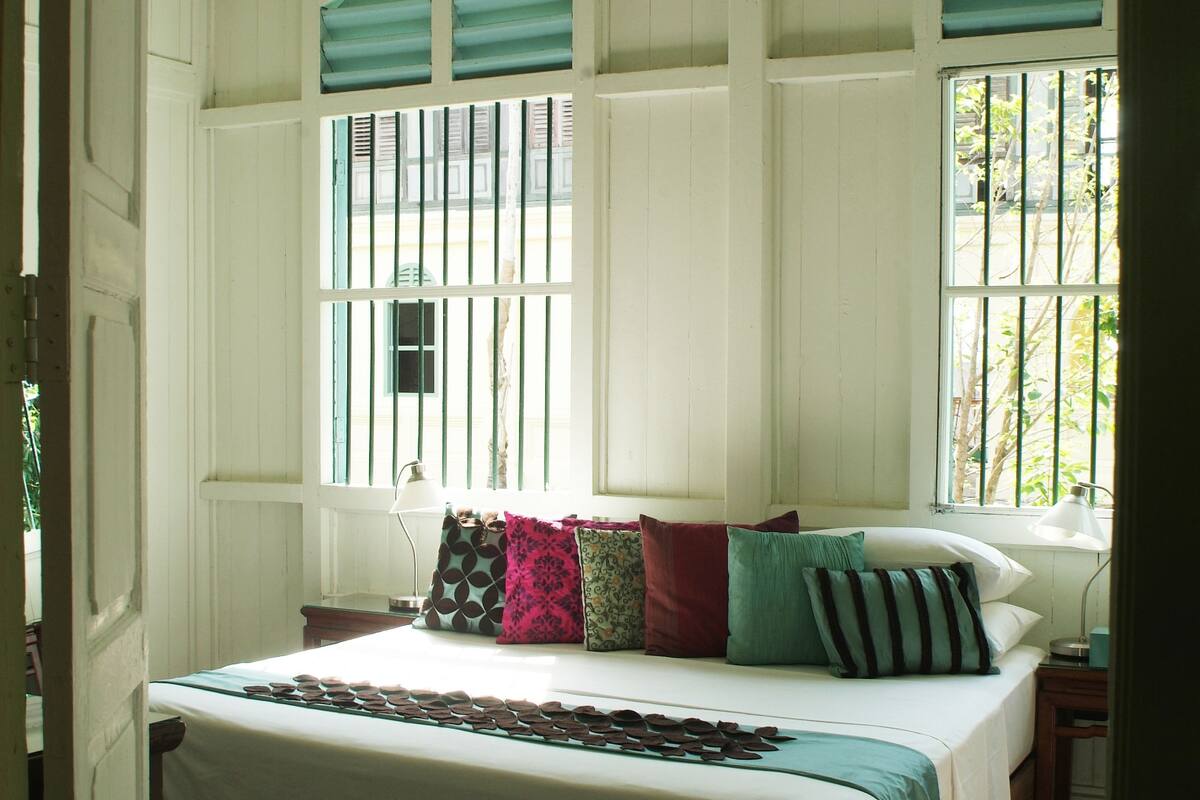 A bedroom with a large bed, two windows with bars, a ceiling fan, and a wooden floor.