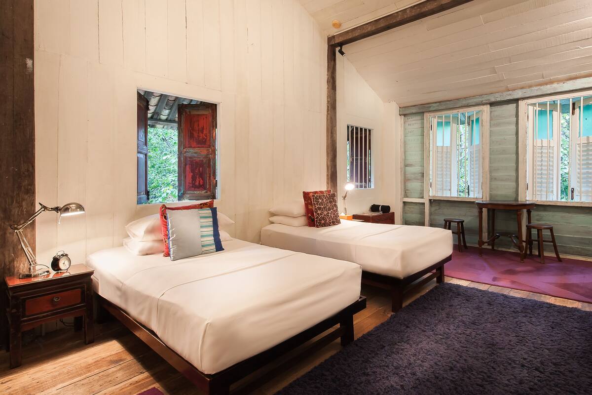 A room with two beds, a nightstand with a lamp, a small table, and a window with bars.