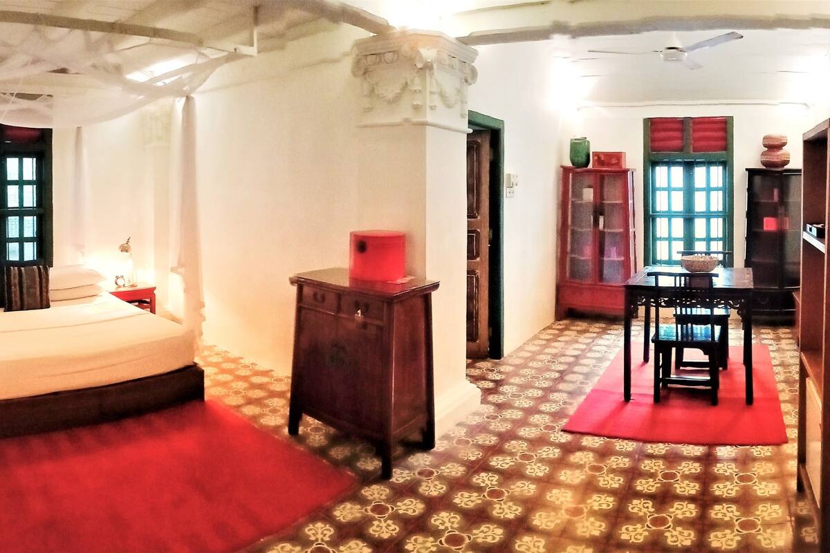 A room with a bed, a desk, and a chair. There is a window with green shutters and a red curtain.