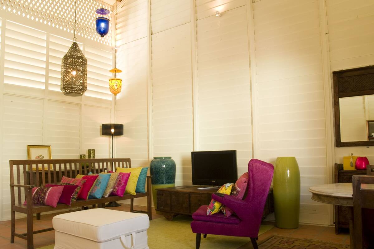 A living room with a wooden bench, a white ottoman, a purple armchair, a green vase, and a television.