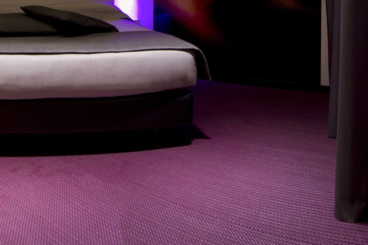 A modern hotel room with a bed, a large wall mural, and purple carpeting.