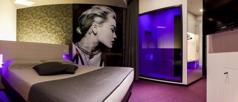 Una habitación de hotel moderna con una cama grande, un baño con cabina de ducha de vidrio y un televisor de pantalla plana.