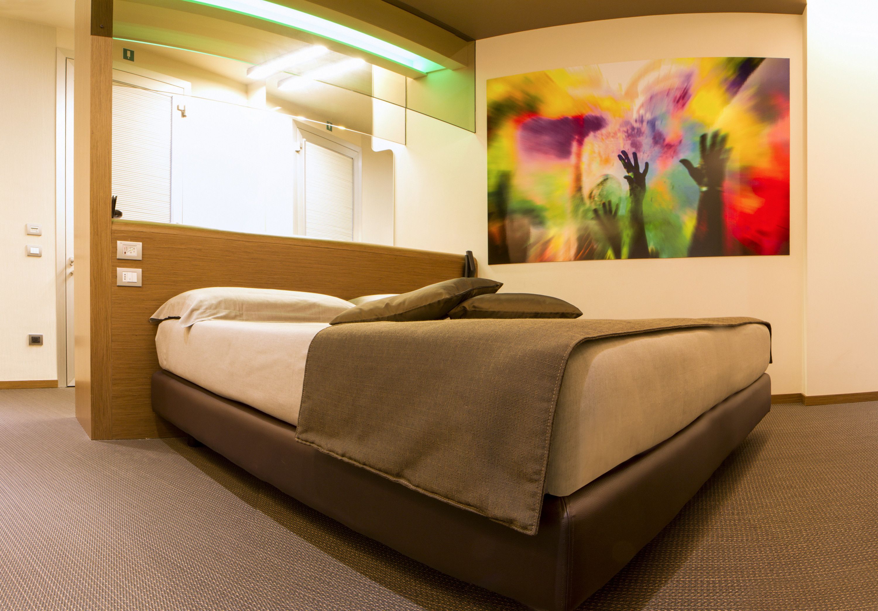 A hotel room with a bed, a wall-mounted TV, and a colorful abstract painting.