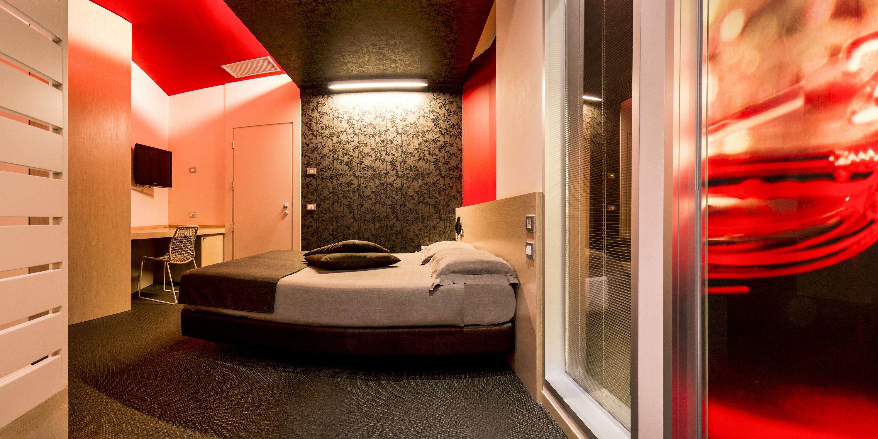 A modern hotel room with a curved bed, a desk, and a large window with blinds.