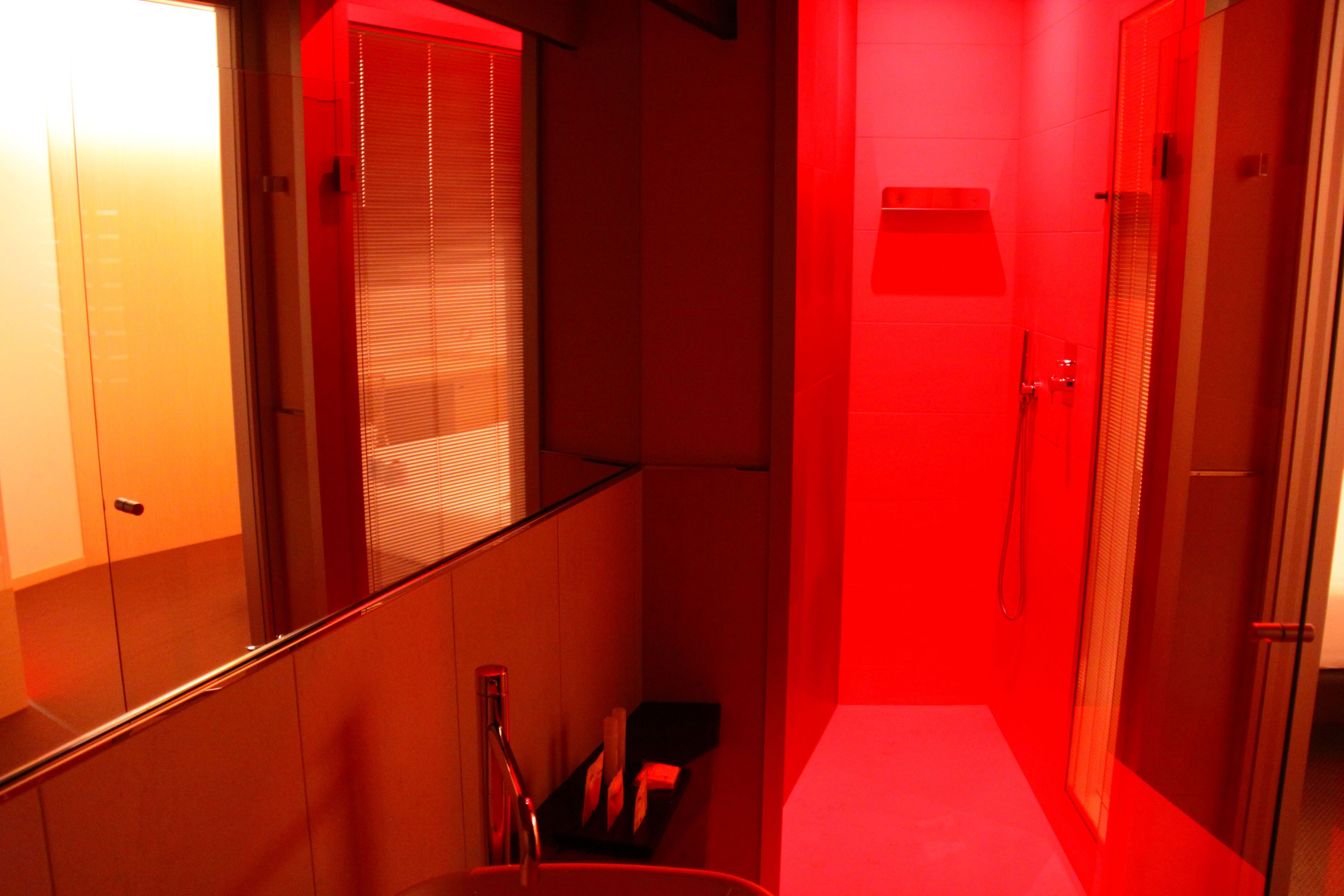 Um corredor estreito com área de chuveiro iluminada em vermelho, uma porta de vidro e um pequeno espaço para sentar.