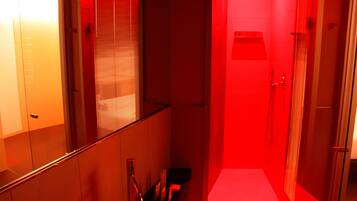 Um corredor estreito com área de chuveiro iluminada em vermelho, uma porta de vidro e um pequeno espaço para sentar.