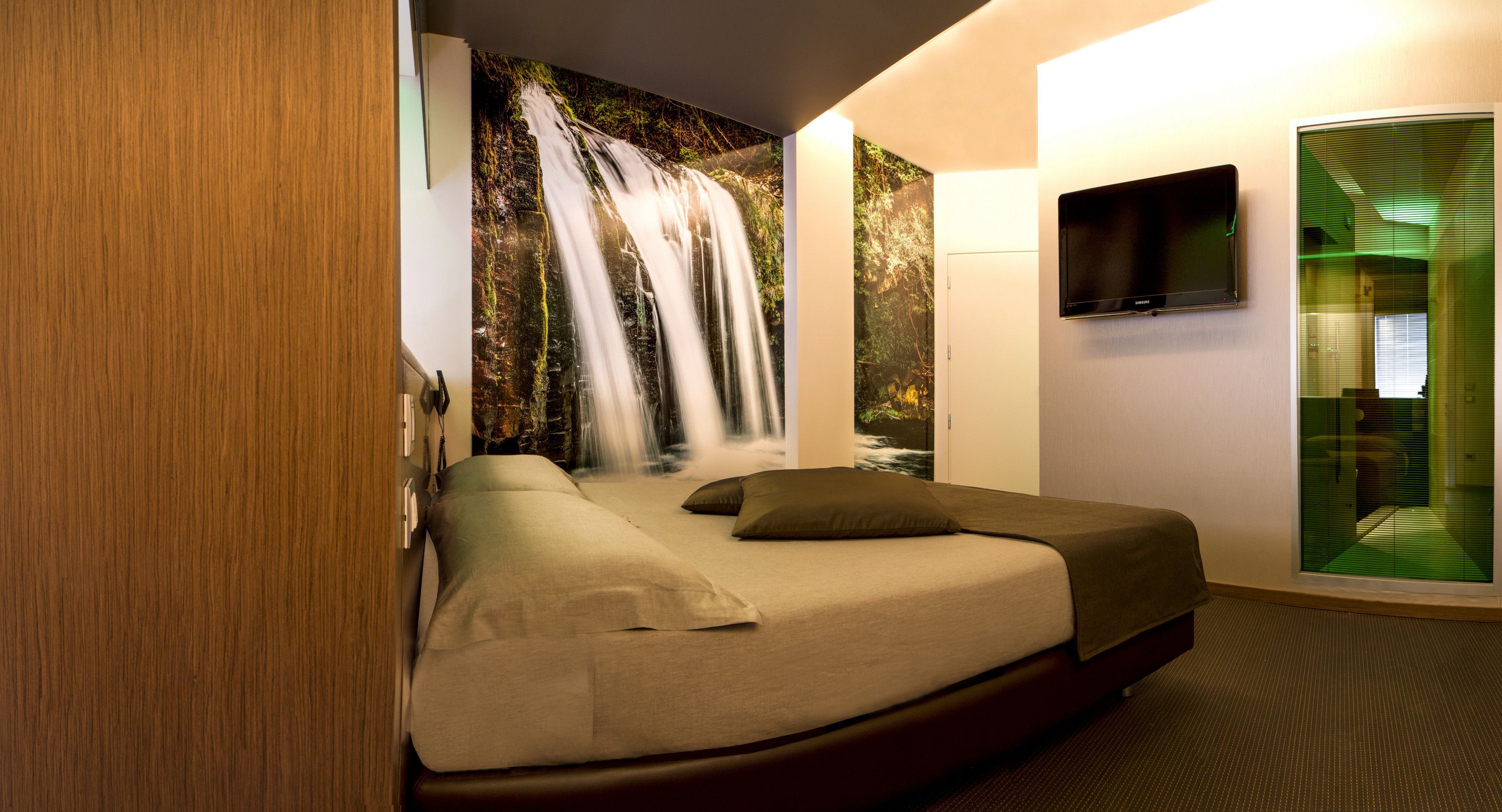 A hotel room with a bed, a TV, and a wall mural of a waterfall.