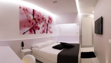 Un dormitorio moderno con un televisor grande montado en la pared, una cama con ropa de cama blanca y almohadas negras, una mesita de noche y una obra de arte en la pared.