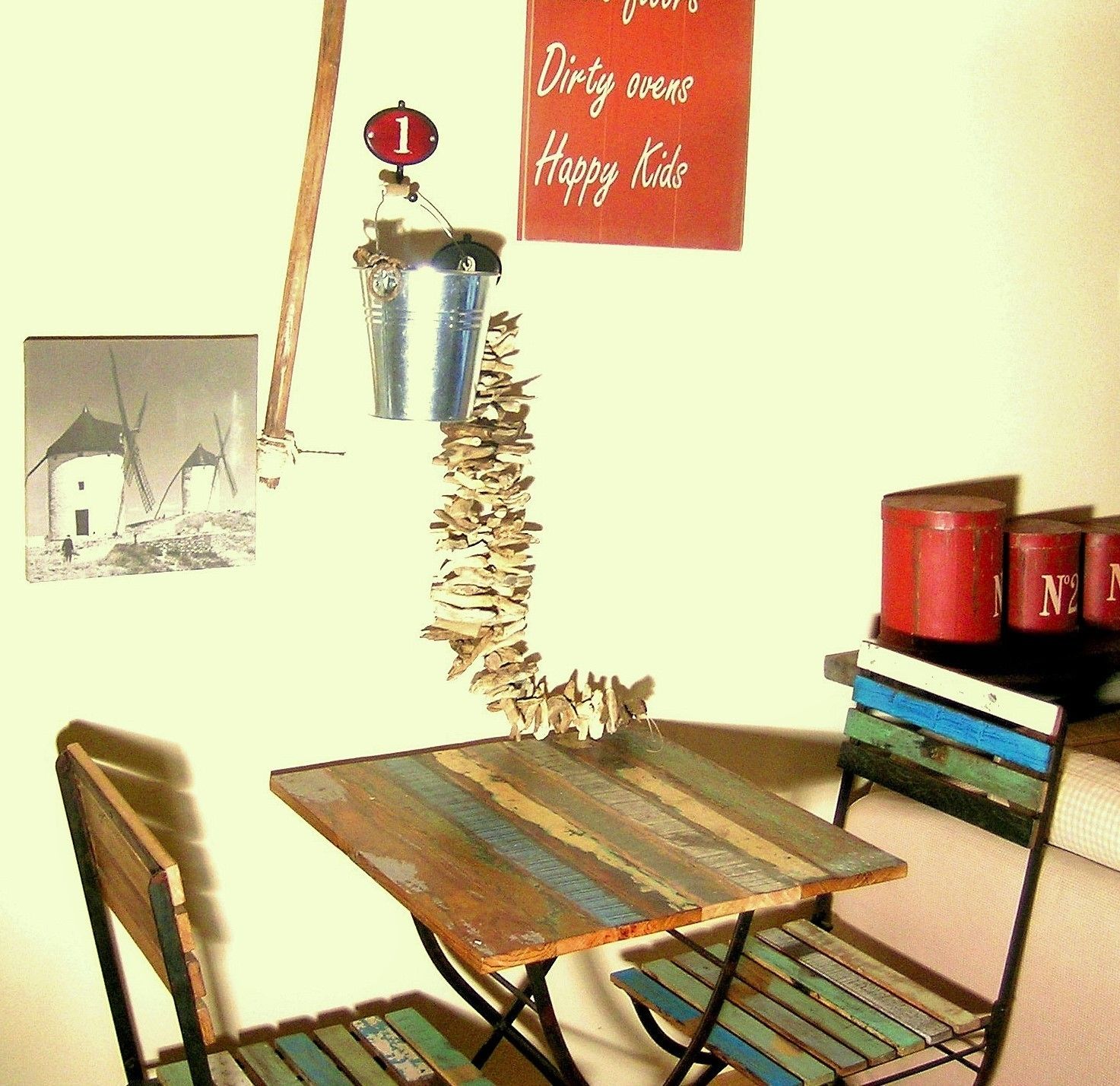A rustic room with a wooden table and chairs, a windmill picture, a metal bucket, and a sign that reads "Dirty ovens Happy Kids".