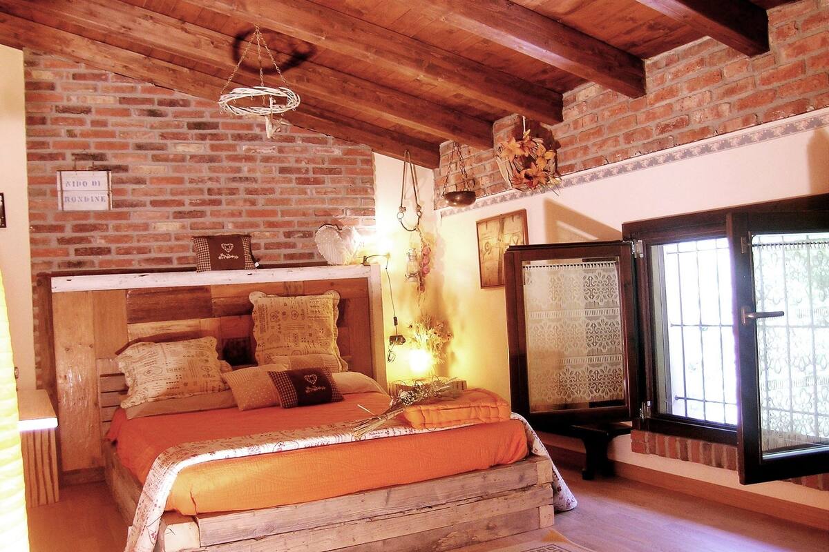 A rustic bedroom with a brick wall, wooden beams, a bed with patterned pillows, a hanging light fixture, and a window with a decorative screen.