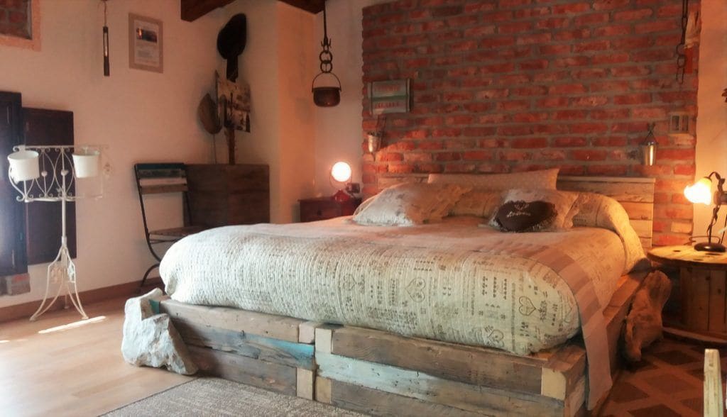 Camera rustica con parete in mattoni, struttura letto in legno e comodino con lampada.
