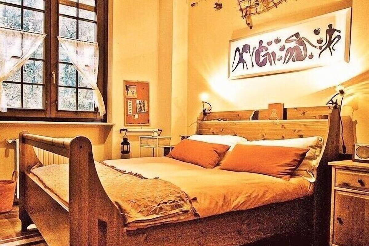 A traditional wooden bed with a headboard, a bedside table, and a nightstand. There is a window with curtains, a wall clock, and a framed artwork above the bed.
