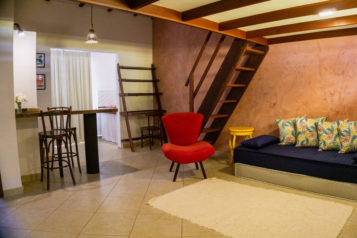 Un espacio de estar acogedor con comedor, una silla roja, un sofá con almohadas tropicales y una escalera de madera.