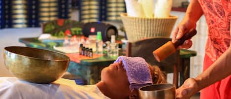 A person receiving a head massage with a bowl and a mallet, in a room with a patterned wall and a basket of bread.