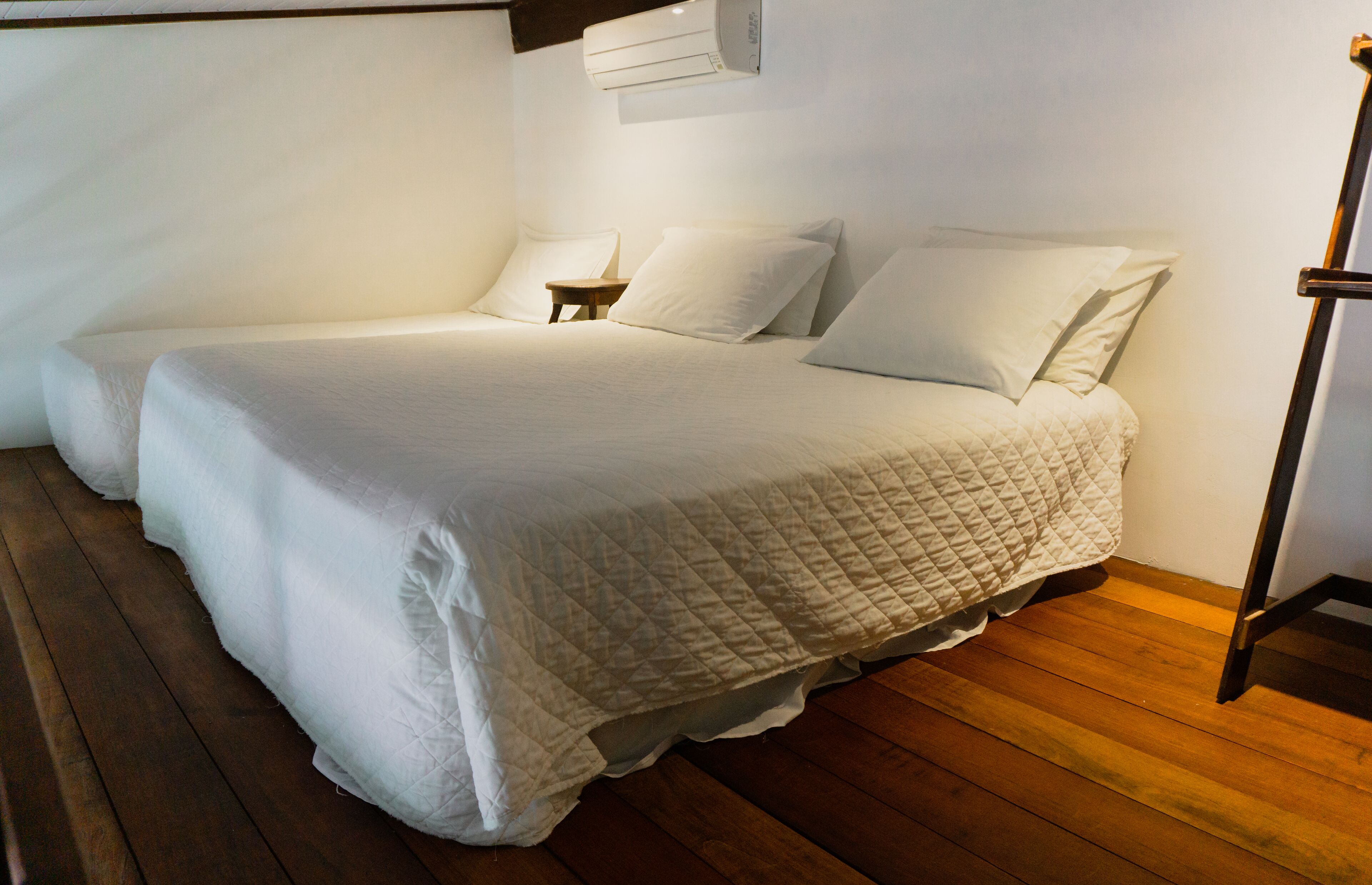 A single bed with a quilted white duvet, a wooden nightstand, and a wall-mounted air conditioner.