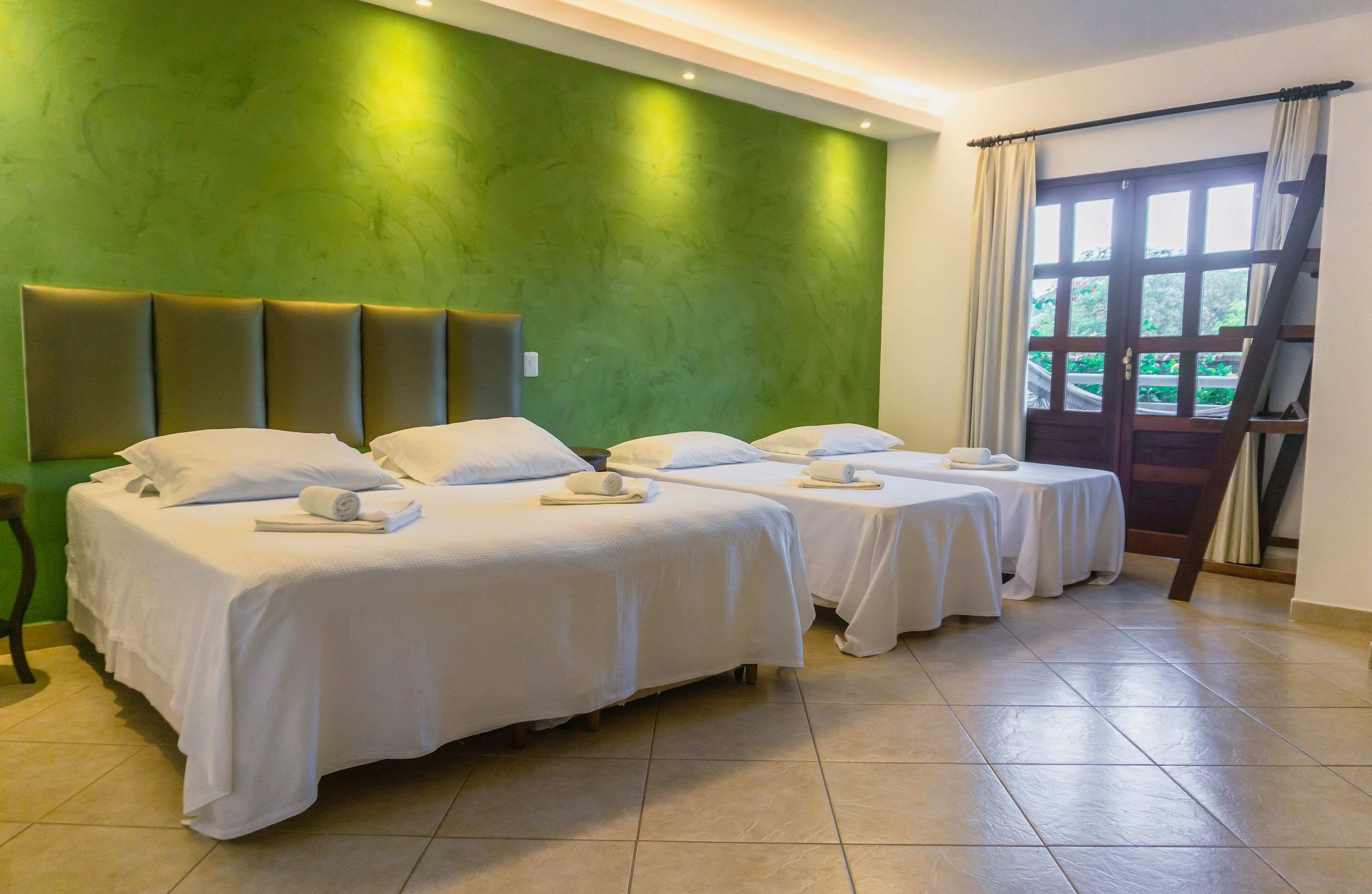 Habitación de hotel con dos camas, una pared de acento verde, una ventana con cortinas y una escalera de madera.