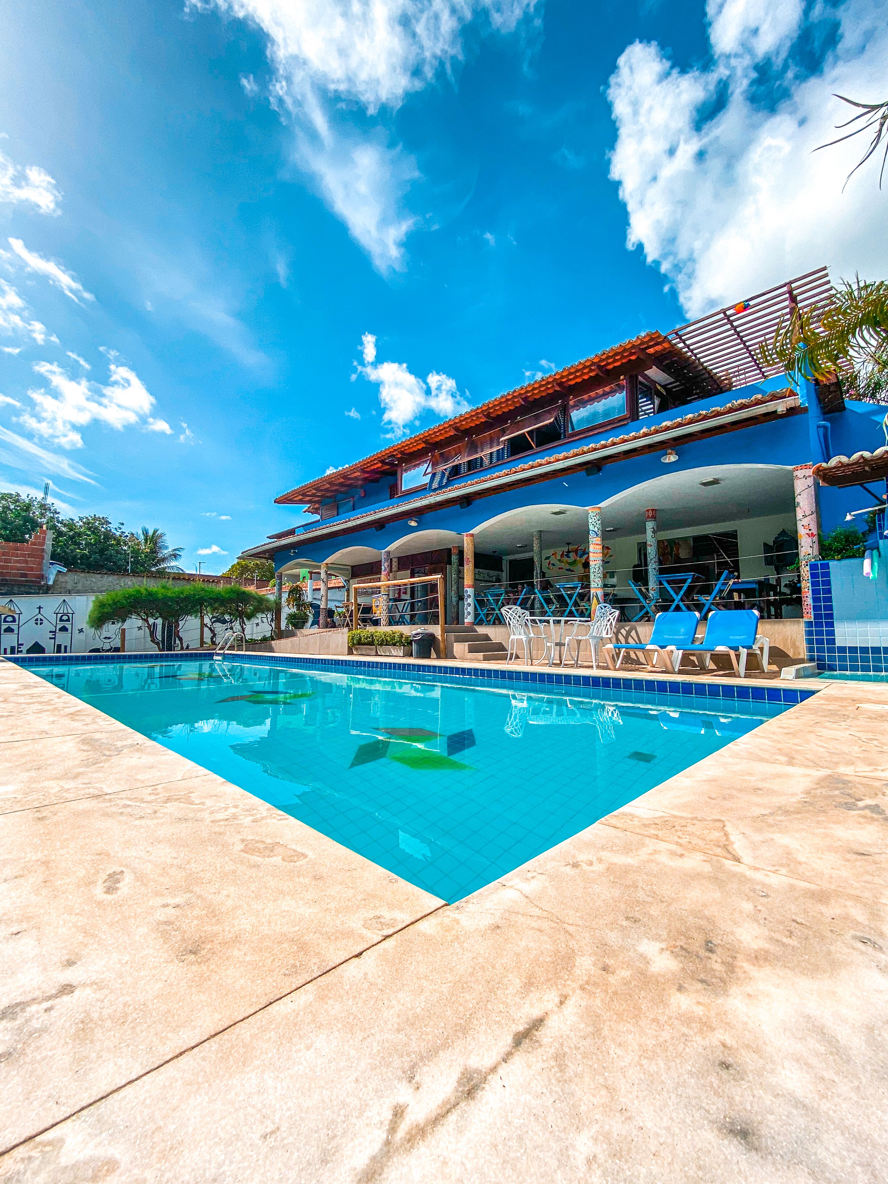 A swimming pool with a tiled deck, a covered patio area with chairs, and a building with a blue facade and red roof.