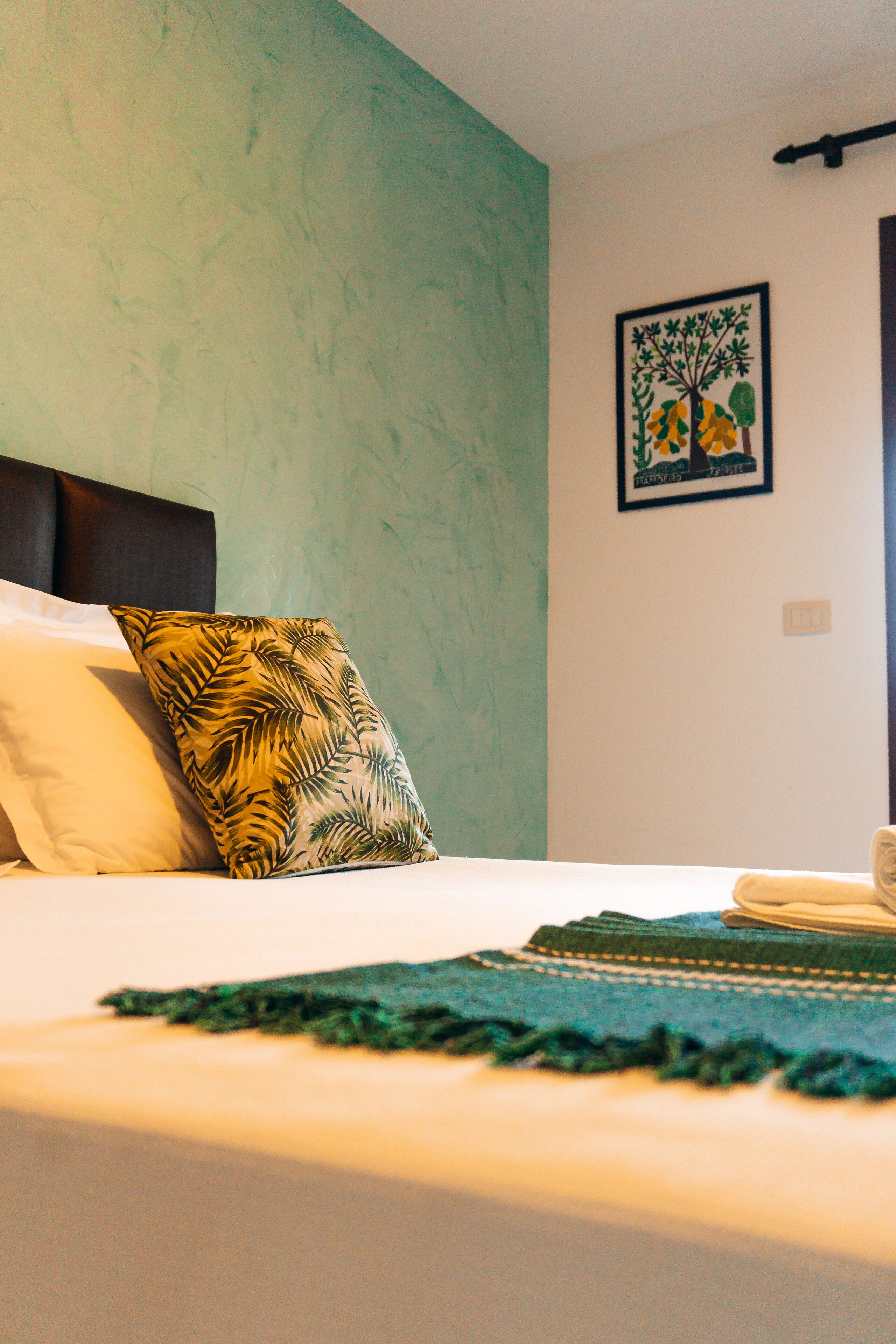 A neatly made bed with a green throw blanket, a pillow with a tropical leaf pattern, and a framed picture of a tree on the wall.