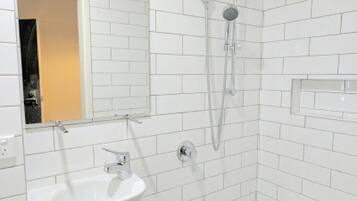 Un baño con paredes de azulejos blancos, un lavamanos blanco y una ducha con cabezal de ducha manual.