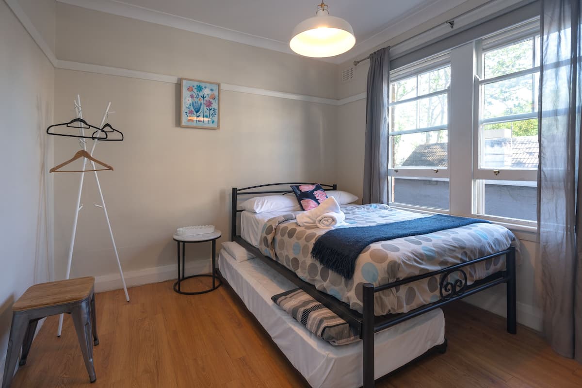 Un dormitorio con cama, una mesita, una silla, un perchero y una ventana con cortinas.