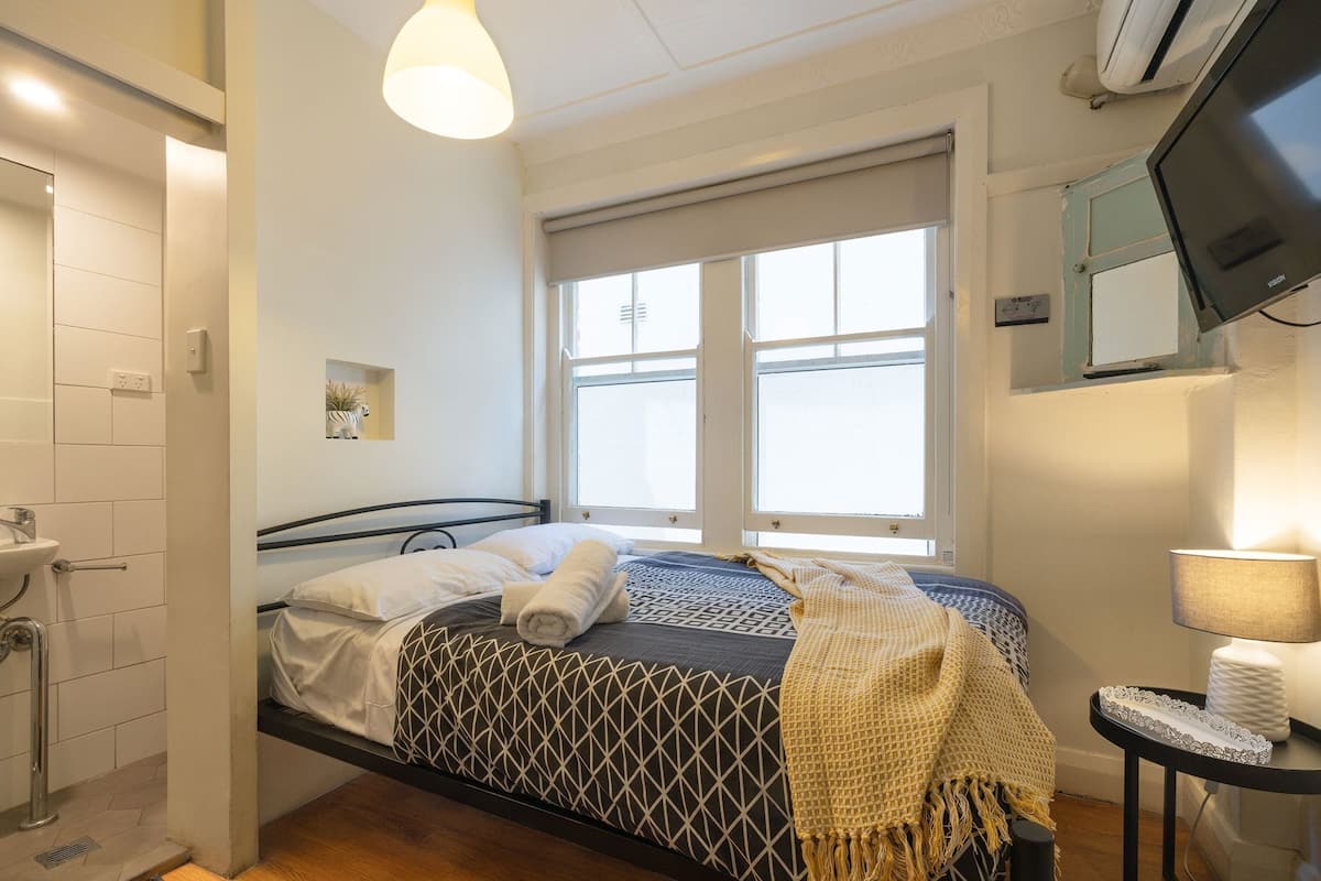 Un dormitorio con cama, ventana, televisor y baño con lavamanos y ducha.