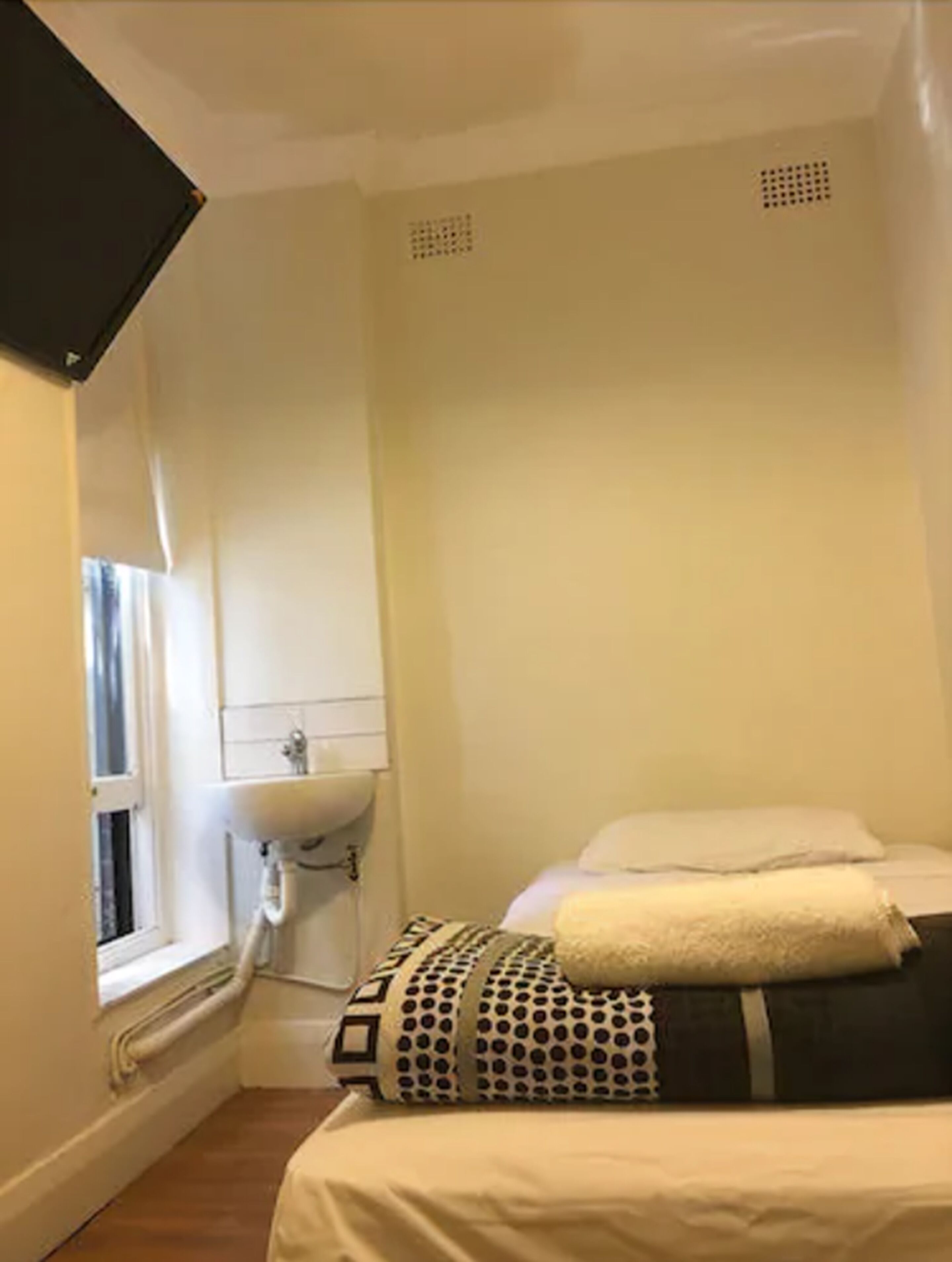 A small, single-bed room with a window, a sink, and a ceiling vent.