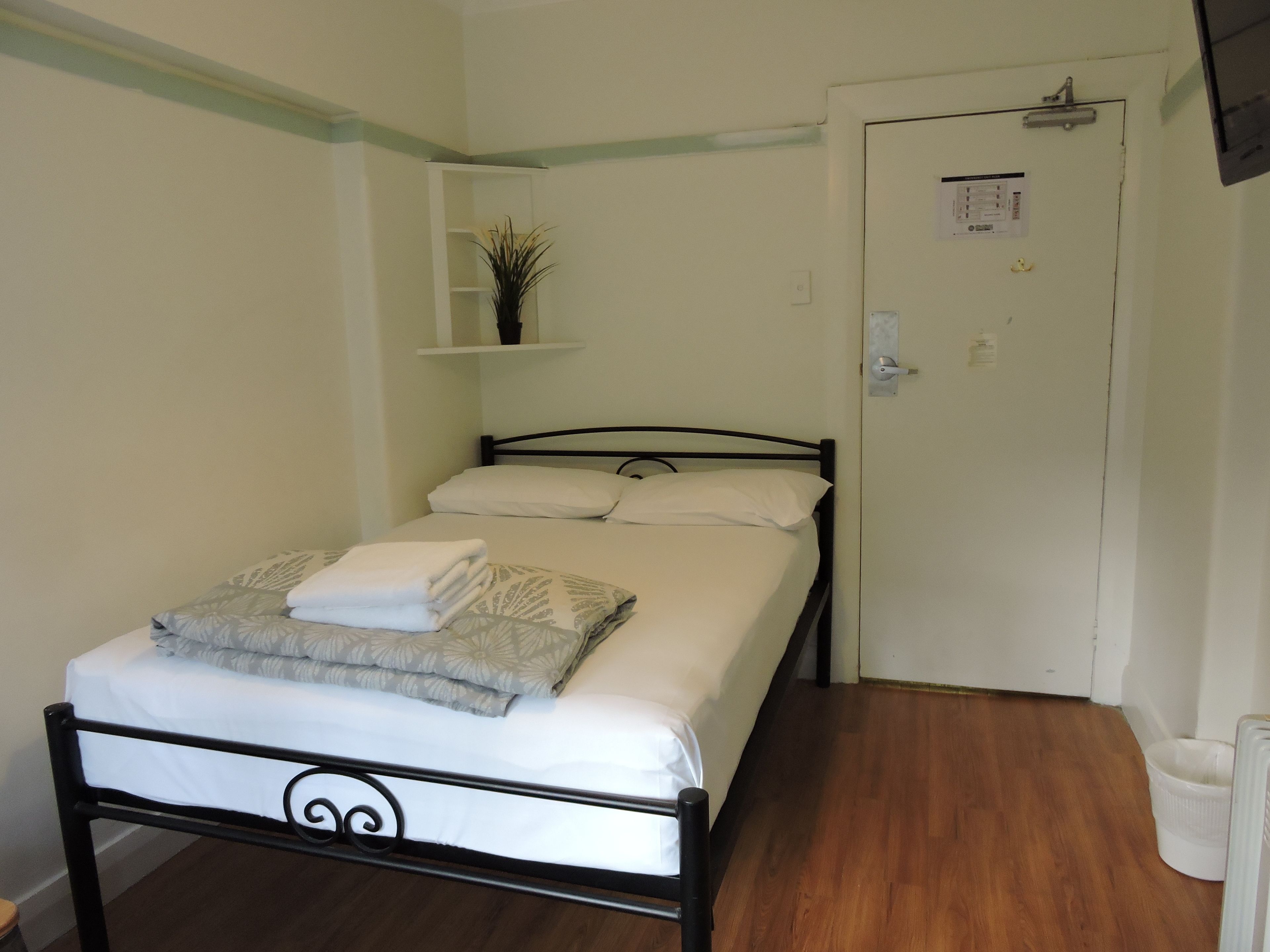 Una cama bien hecha con ropa blanca y una toalla doblada, cabecera de metal negro, piso de madera y una puerta blanca con un tablón de anuncios pequeño.