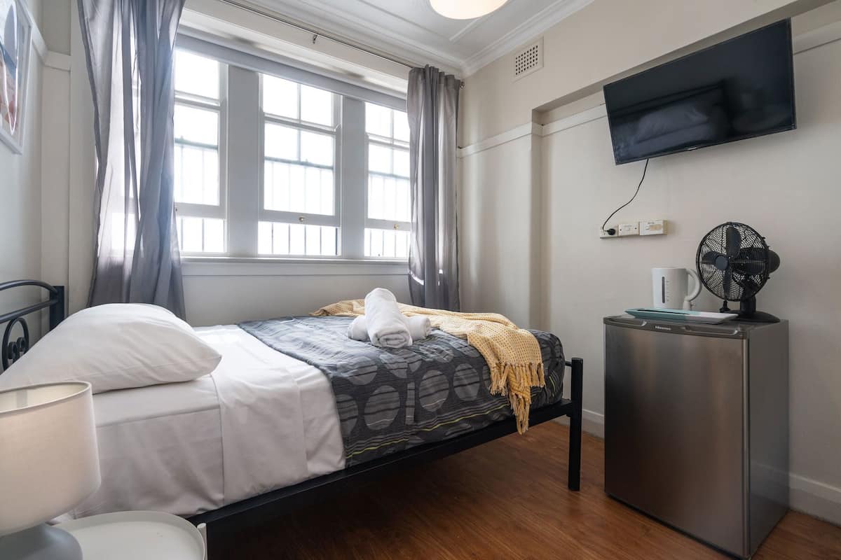 Un dormitorio con cama, escritorio, televisor montado en la pared y ventana con cortinas.