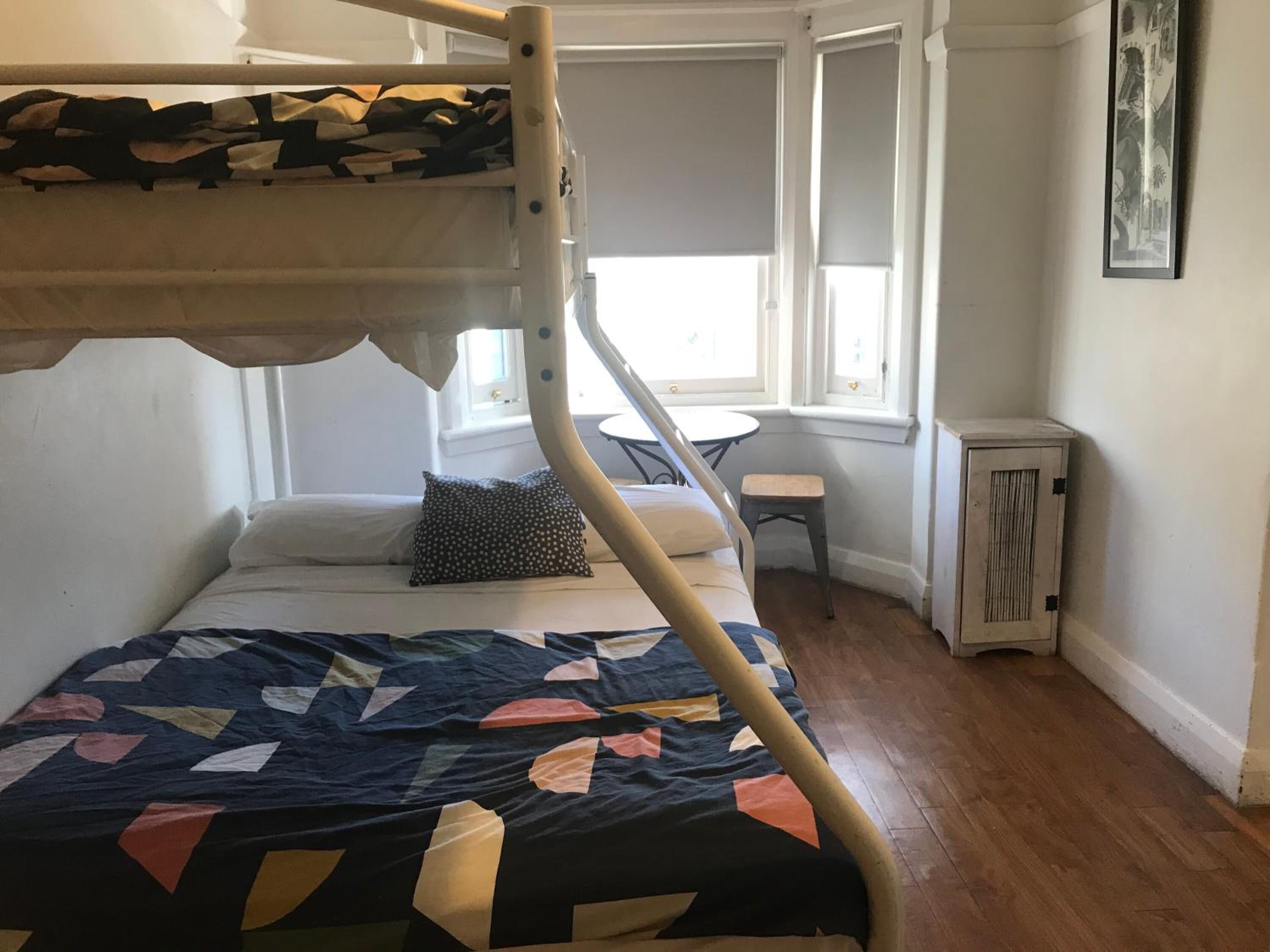 A bunk bed with a patterned quilt, a small table, and a chair in a room with wooden flooring and a window.