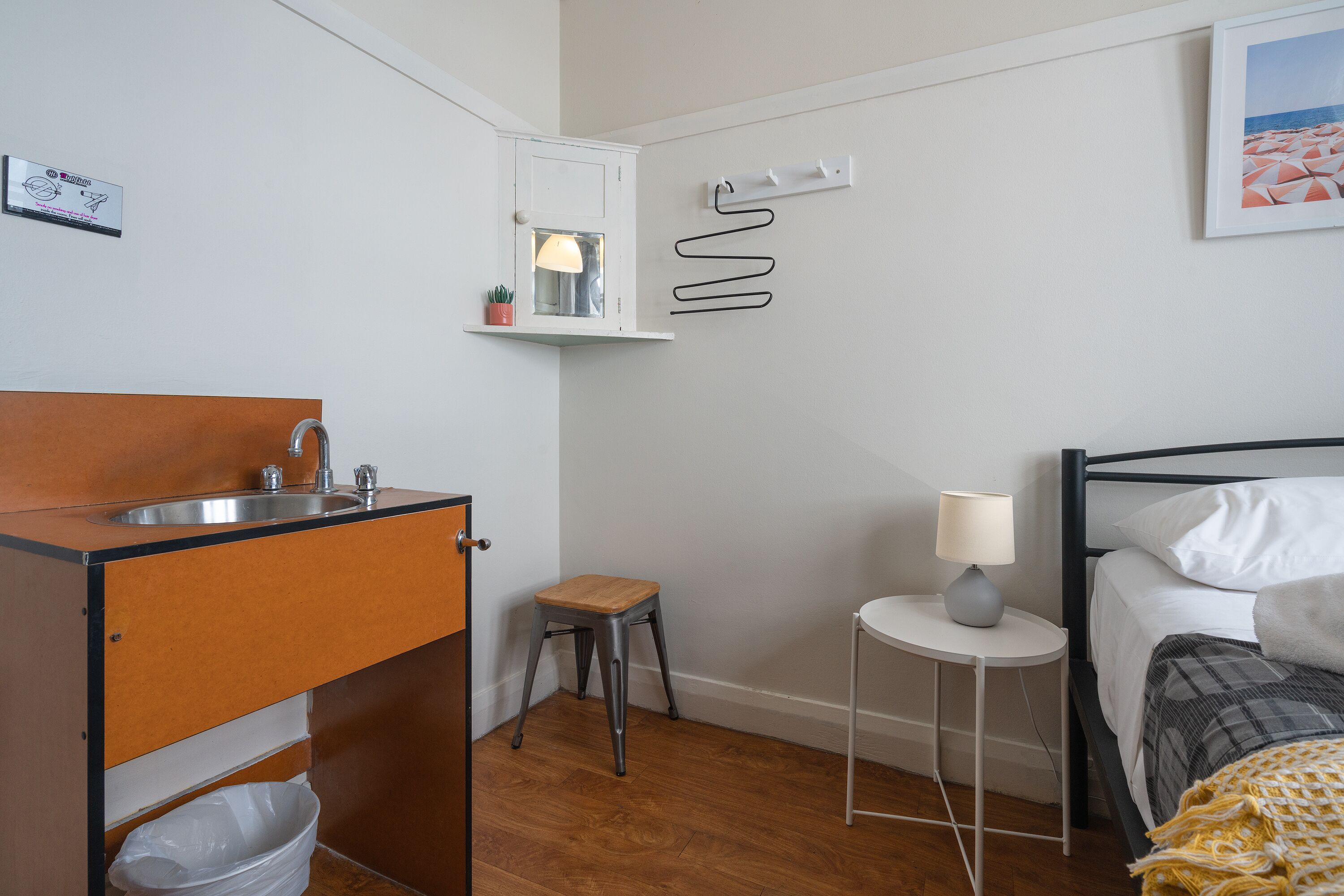 A compact bedroom with a wooden floor, a bed with white bedding, a bedside table with a lamp, a small stool, a cabinet with a sink, and a wall-mounted shelf with a mirror.