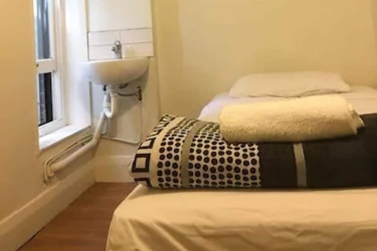 Una habitación pequeña con cama individual, ventana, lavamanos y rejilla de ventilación en el techo.