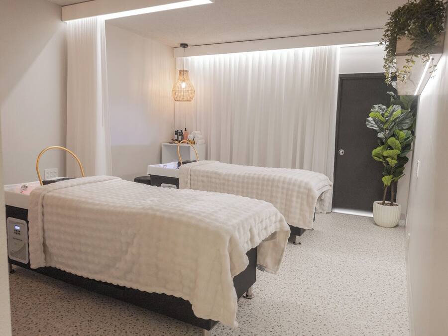 A hotel room with two beds, a bedside table, a lamp, a potted plant, and a door.