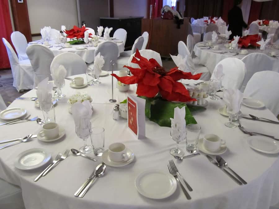 Uma sala de banquetes com mesas redondas montadas para um evento formal, com toalhas brancas, talheres e centros de mesa com flores de poinsétia vermelhas.