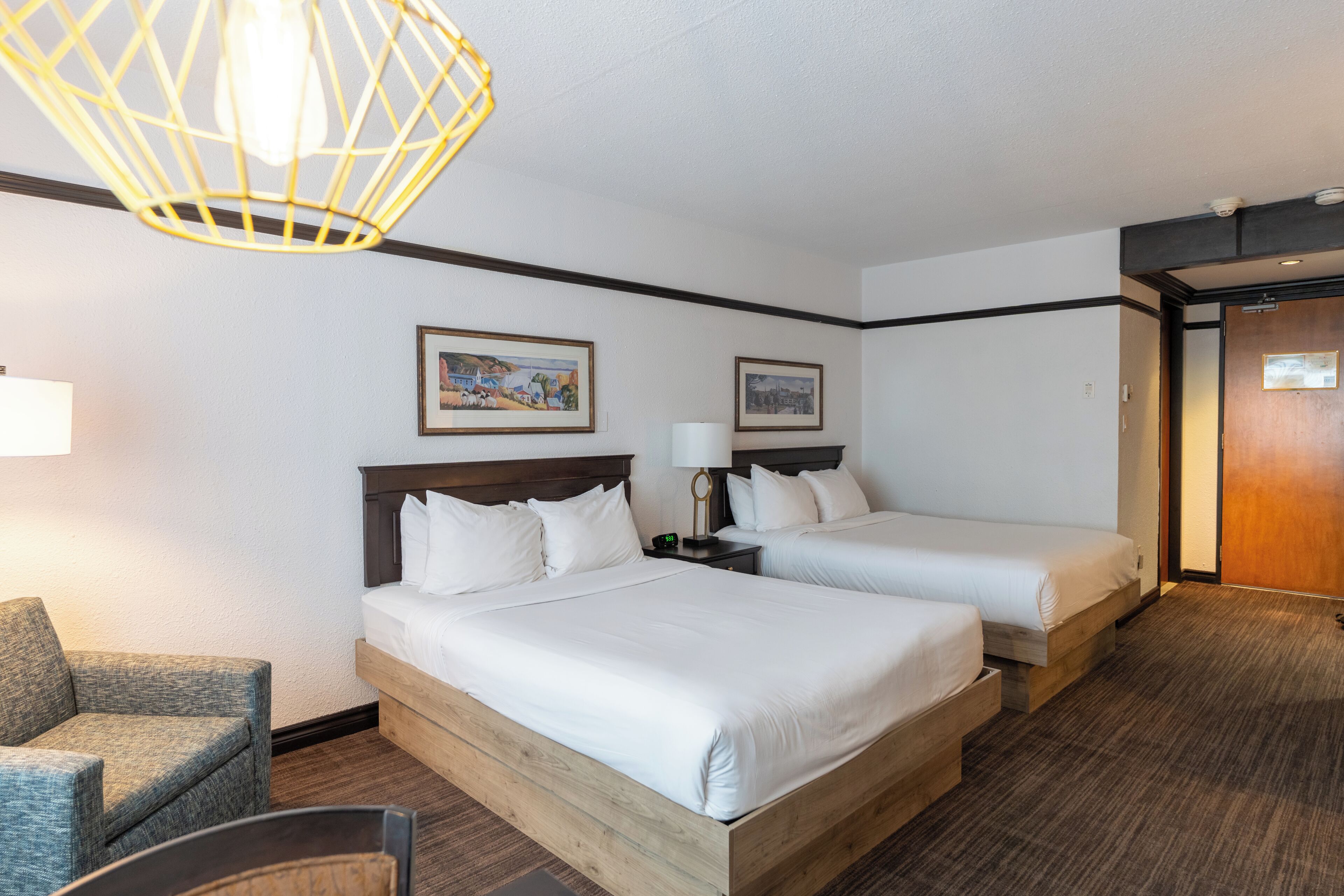 A hotel room with two beds, a chair, a lamp, and two framed pictures on the wall.