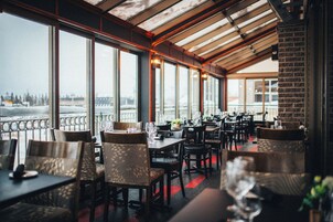 Un restaurante con vistas a un paisaje nevado a través de amplios ventanales. Las mesas están dispuestas con sillas, y el interior presenta un diseño moderno con techo de madera.