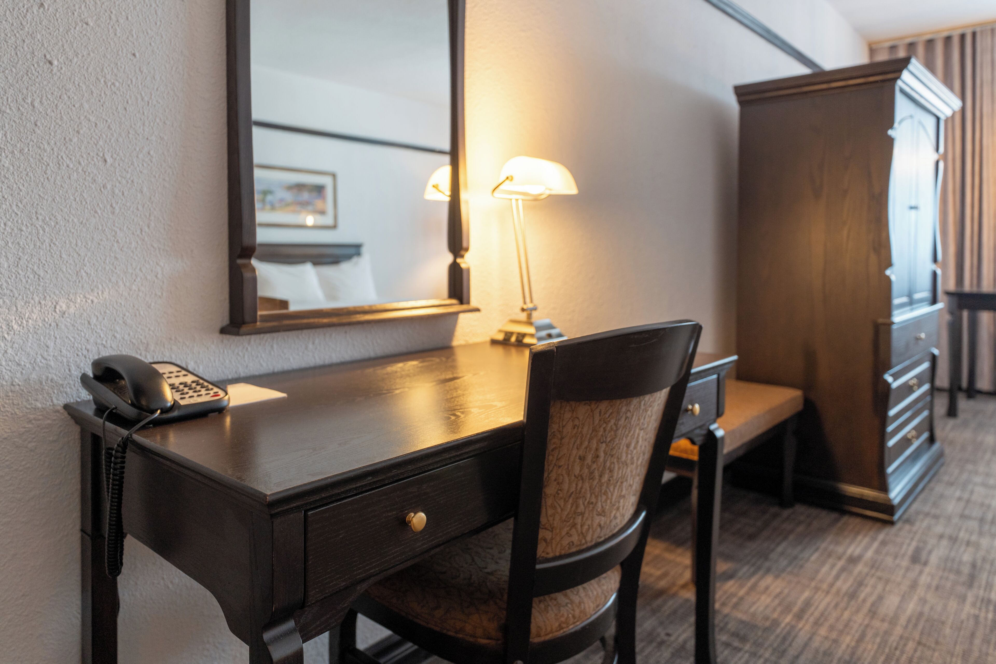 Une chambre d’hôtel avec un bureau en bois, un téléphone, une lampe, une chaise et une armoire.