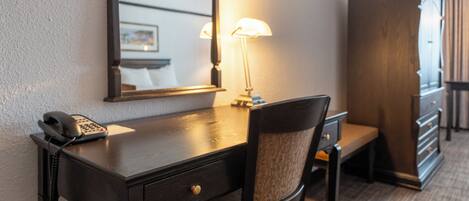 A hotel room with a wooden desk, a telephone, a lamp, a chair, and a wardrobe.