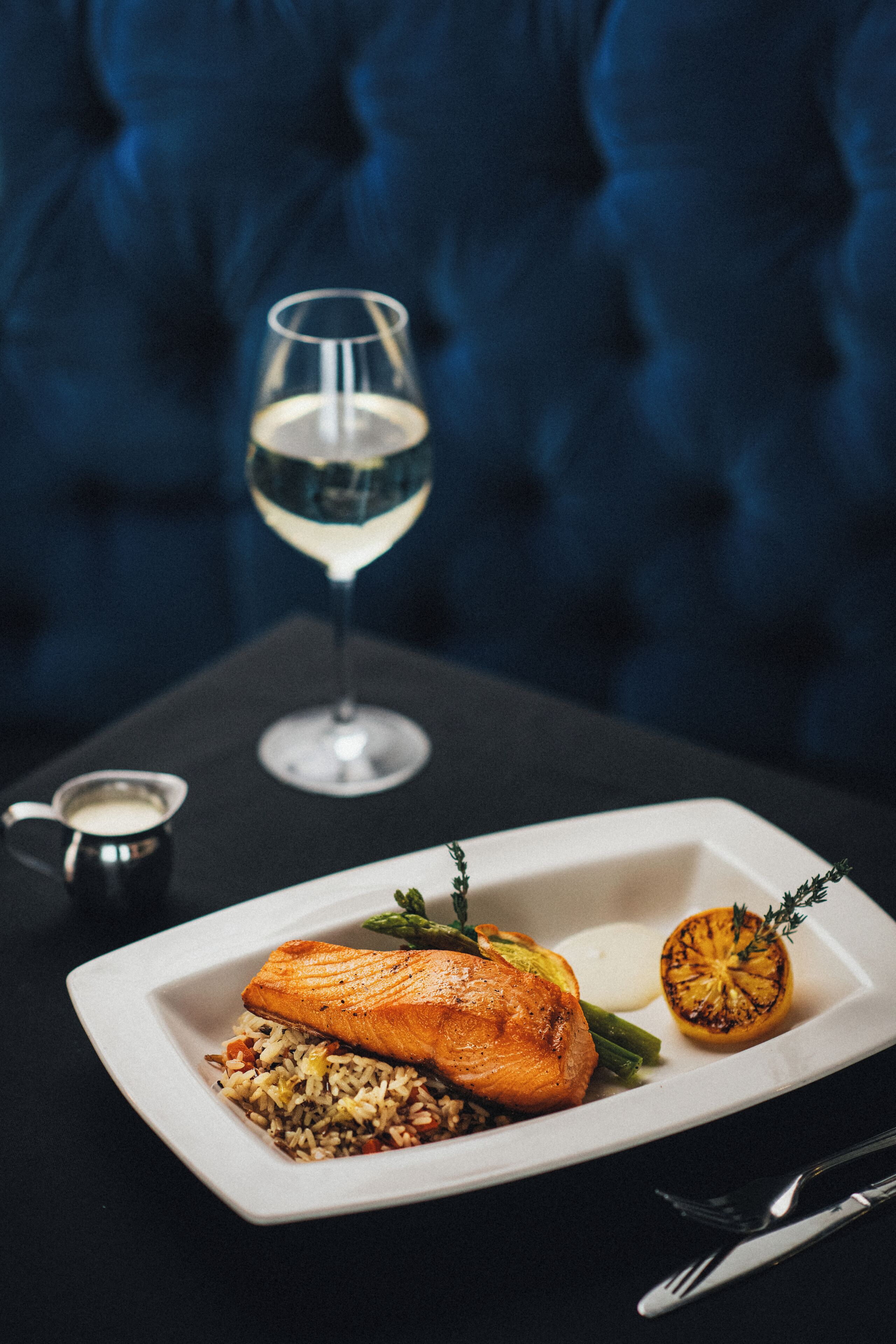 A plate of food with a glass of white wine, a small bowl of sauce, and a knife and fork on a table.