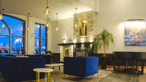 A modern lounge area with a large window, seating, a piano, and a fireplace. - Le Montagnais (Saguenay)
