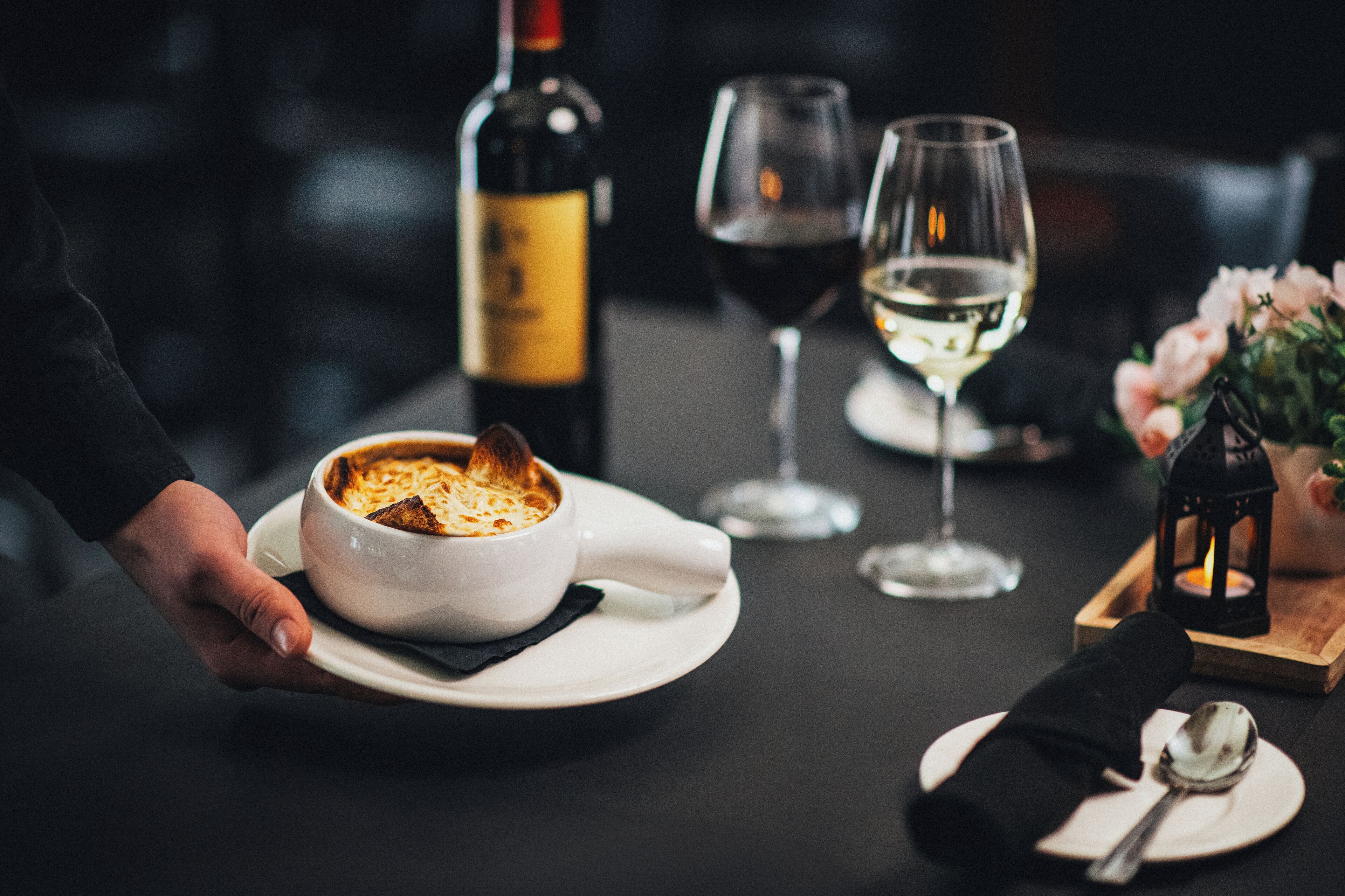 Una persona sirve un plato de comida, una botella de vino y copas sobre una mesa con vela y flores.