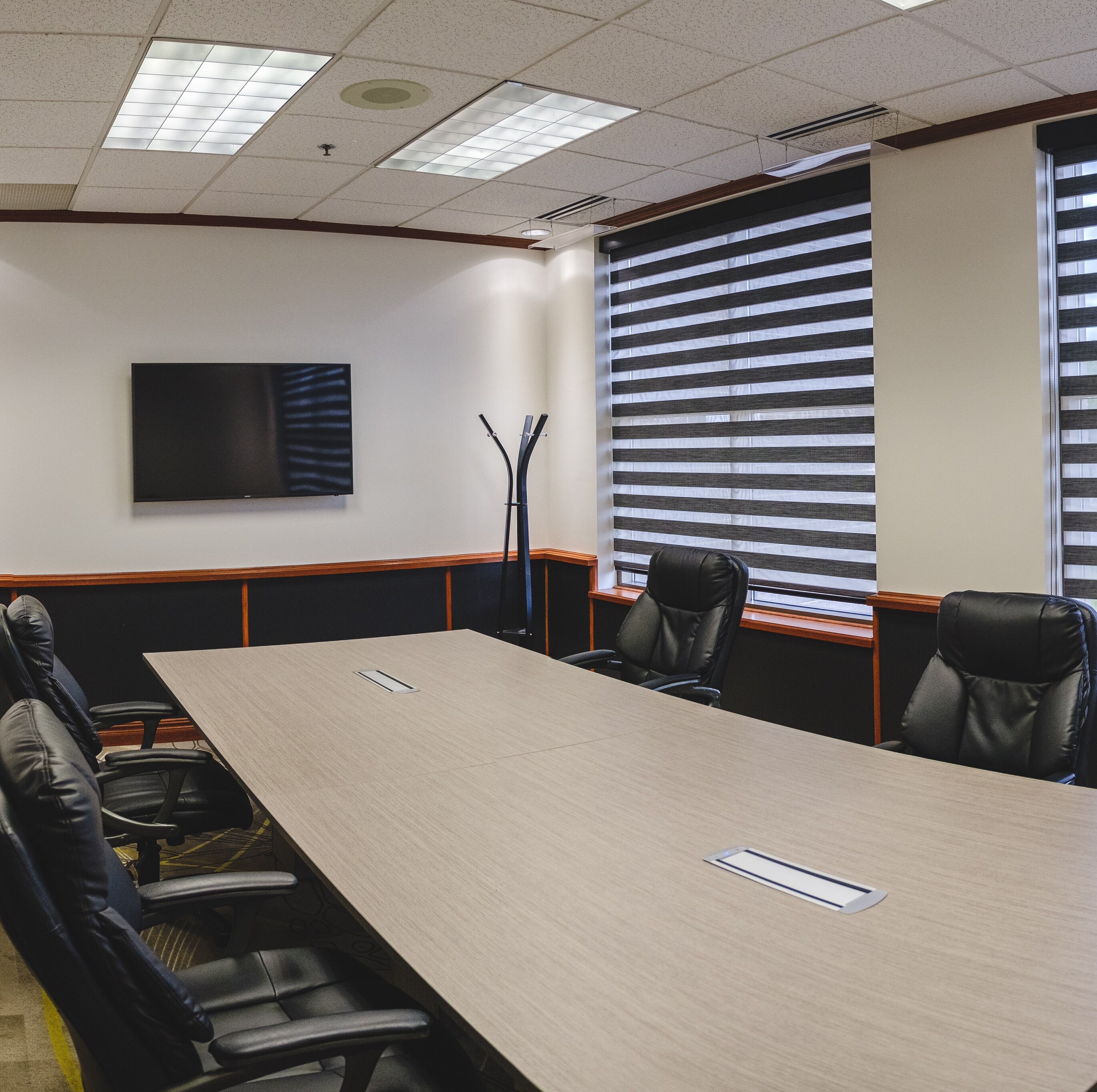 A conference room with a long table, four chairs, a flat-screen TV, and a window with blinds.