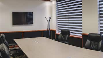 A conference room with a long table, four chairs, a flat-screen TV, and a window with blinds.