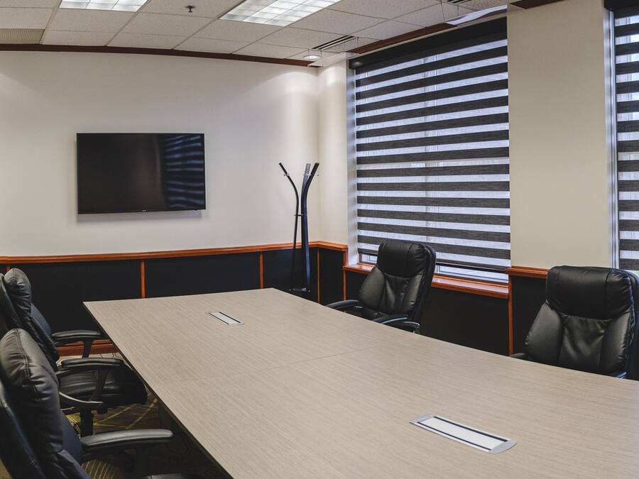 A conference room with a long table, four chairs, a flat-screen TV, and a window with blinds.