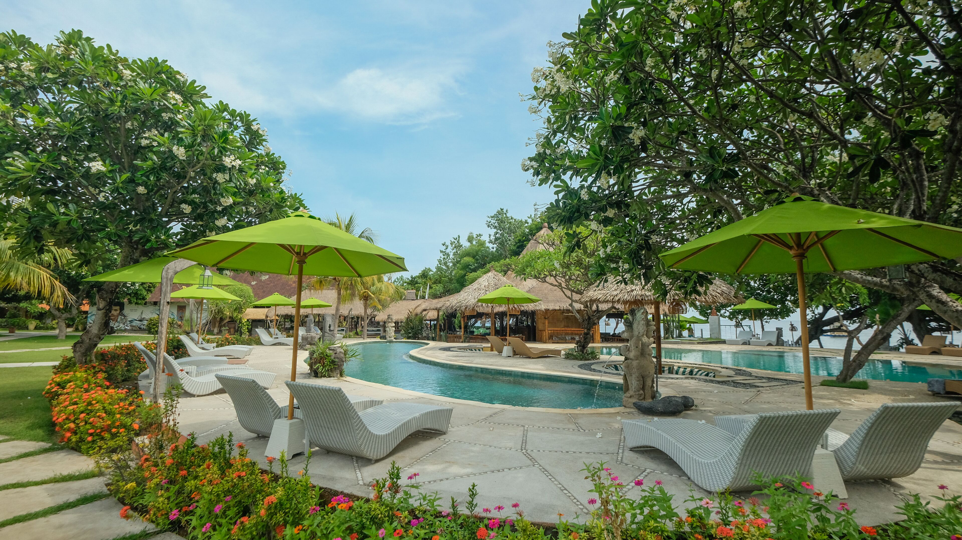 Zona junto a la piscina con sillones de descanso y sombrillas, rodeada de exuberante vegetación y un cielo despejado.