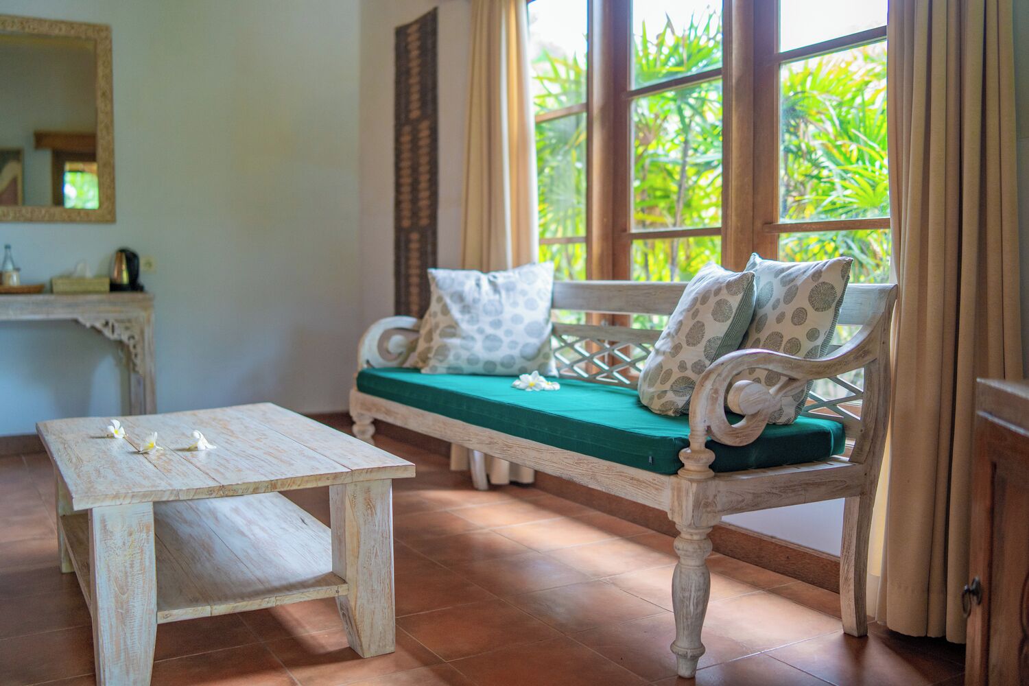 Un banc en bois avec un coussin vert et des oreillers à motifs, une petite table ornée de fleurs blanches et une fenêtre donnant sur un paysage verdoyant.