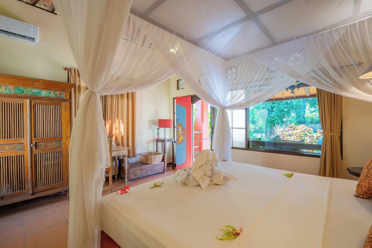 A hotel room with a bed, a wooden wardrobe, a window with a view of greenery, and a mosquito net.