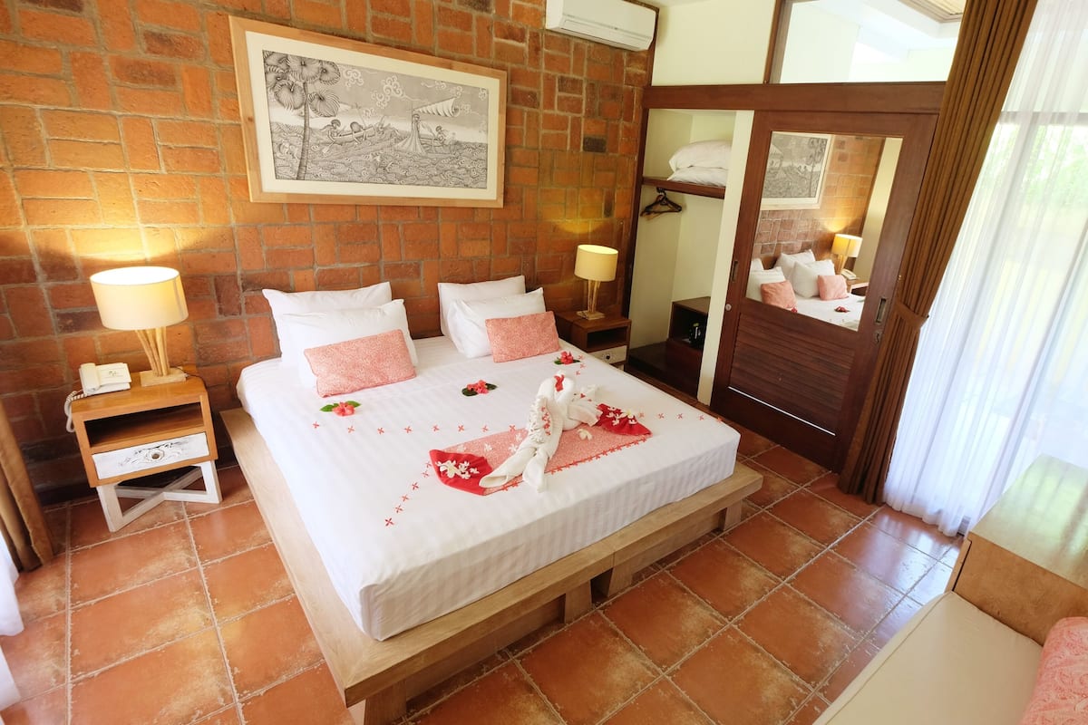 A hotel room with a brick wall, a bed with white and pink bedding, a bedside table with a lamp, a wooden bench, a TV, and a mirror.