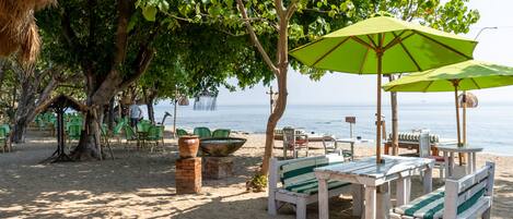 A beachside seating area with tables and chairs, a large umbrella, and a view of the sea.