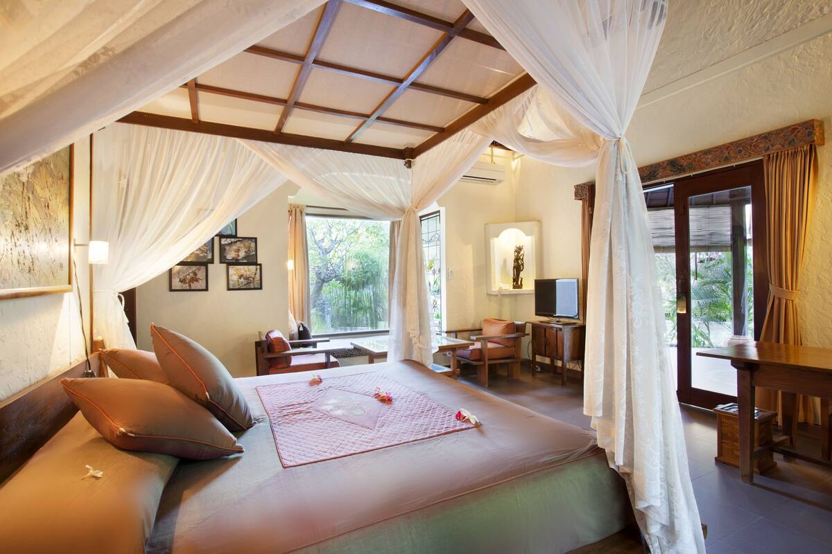 A bedroom with a bed, pillows, a bedside table, a desk, a chair, and a window with curtains.