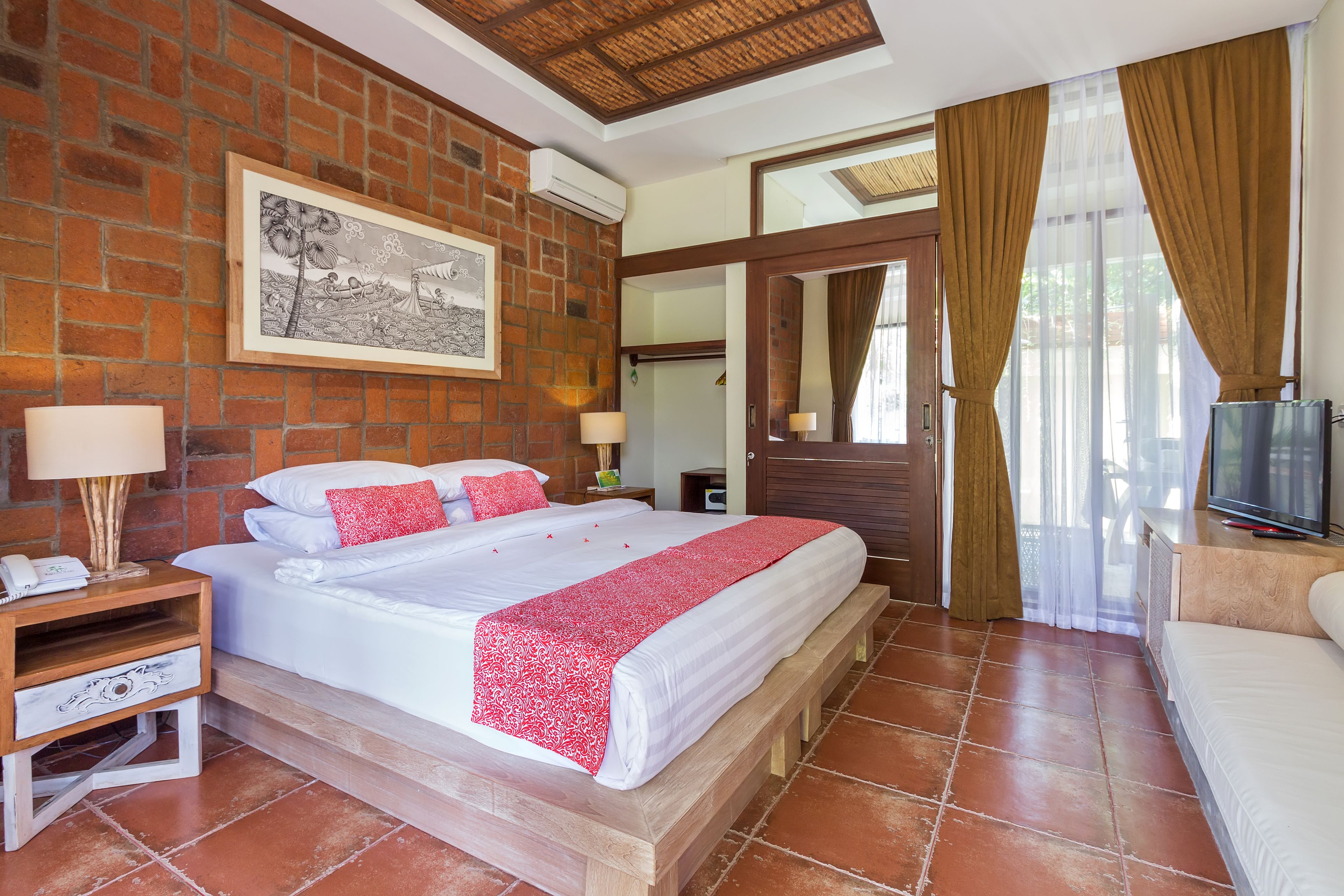 A hotel room with a brick wall, a large bed with white bedding and a red throw, a wooden bench, a nightstand with a lamp, and a TV on a wooden stand.